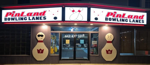 Exterior of PinLand Bowling Lanes at night with bright signs and large bowling pin cutouts flanking the glass entrance.
