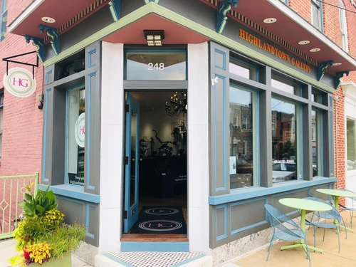 The colorful storefront of Highlandtown Gallery features blue trim, a red awning, and small outdoor café tables on the sidewalk.