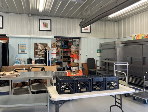 A clean food pantry interior with industrial refrigerators, metal prep tables, and organized food boxes.