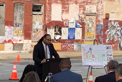 A man speaks at a podium in front of blighted buildings with a neighborhood development site map on an easel.