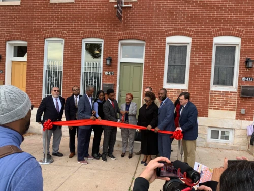 Officials hold a large red ribbon for a ceremonial cutting in front of newly renovated brick rowhouses.