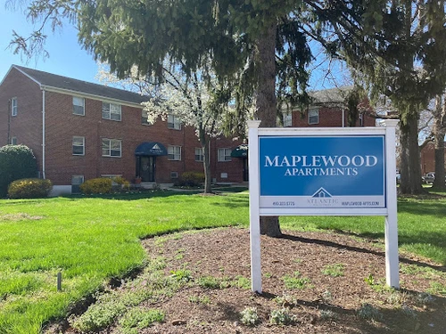 A large blue sign reads "Maplewood Apartments" on a grassy lawn in front of a multi-story brick apartment building.