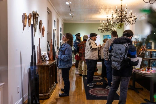 Visitors explore an art gallery with hardwood floors and chandeliers, viewing various sculptures and paintings displayed on walls and tables.