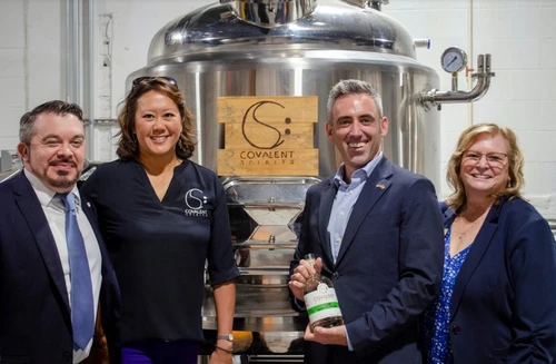 Four people posing in front of a still holding a bottle.
