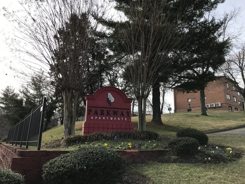 A red arched monument sign for "Parkway Apartments" stands on a grassy hill surrounded by trees, small bushes, and yellow flowers under a clear sky. In the background, a multi-story brick apartment building is visible on the slope.