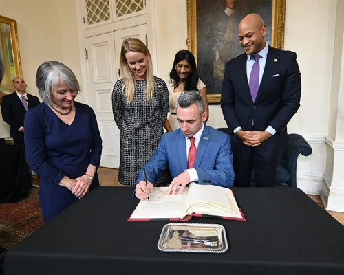 A man signing a document on a table surrounded by people.