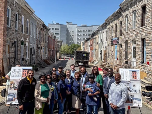 A group of people, including community members and officials, pose for a photo on a city street lined with historic rowhouses. Two large posters displaying architectural renderings of home renovations are visible on easels in the foreground.