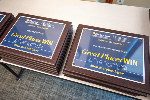 A stack of awards with the awardee's name and Department "Great Places Win" branding sit on a table.