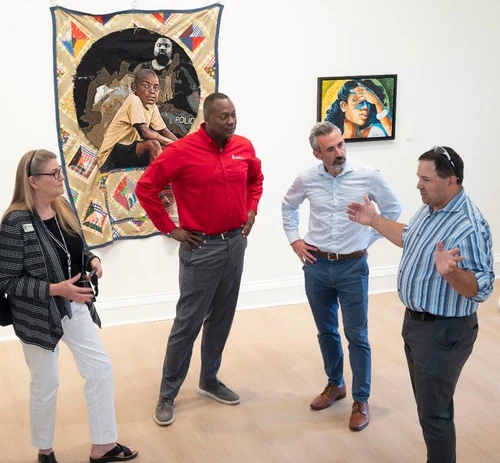 Two men and a woman listen to another man speak. In the background are two painting.