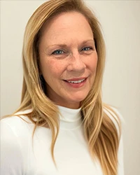 Portrait of a smiling white woman with long blonde hair against white background. Wears white turtleneck top.