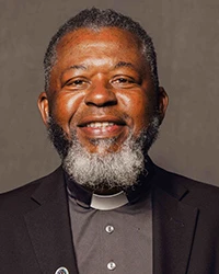 Man with gray hair and beard, dark suit jacket and gray shirt smiling at camera