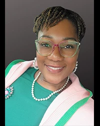 Portrait of a smiling Black woman with braided hair, green glasses, pearl earrings/necklace, pink cardigan over green top with brooch.
