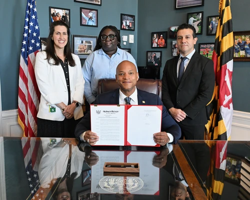 A seated Governor Wes Moore smiles while holding an open red folder displaying a signed government document at a desk in an office. Three staff members stand behind them, with the U.S. flag on one side and the Maryland state flag on the other. Framed photos decorate the wall in the background, and a ceremonial pen rests on the desk in front of the official.