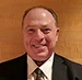 Headshot portrait of Steven Cooper, a man with thinning hair, wearing a suit and tie, smiling against a warm wooden wall.