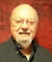 Portrait of Al Sonnenstrahl, an older man with glasses, a gray goatee, and a black striped shirt, smiling against a warm red background.