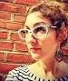 Close-up portrait of Lori Berrong, a woman with curly hair, cat-eye glasses, and statement earrings, looking to the side against a red brick wall.