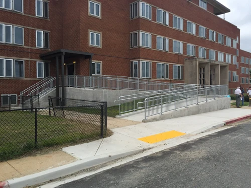An accessible concrete ramp with metal handrails leading to an entrance of a large multi-story red brick building.
