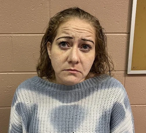 A woman stands against a brick wall wearing a blue and white sweater. She has short brown hair. 