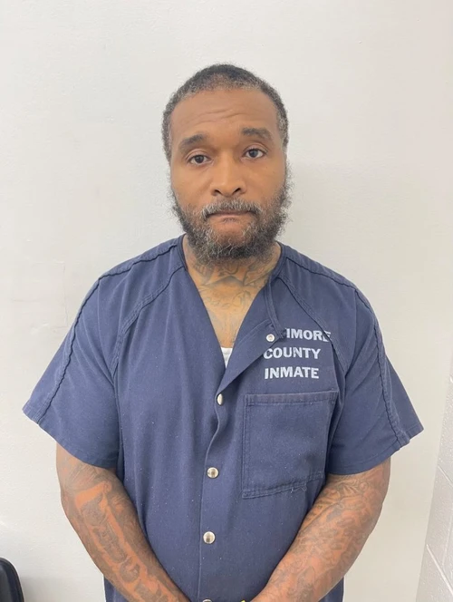 An African American male stands in front of a white wall waering a howard county inmate blue jumpsuit 