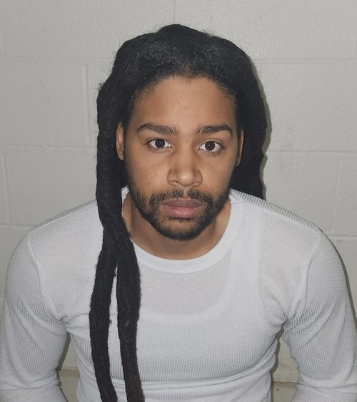 An African American male stands in front of a gray wall wearing a long-sleeved white shirt. 