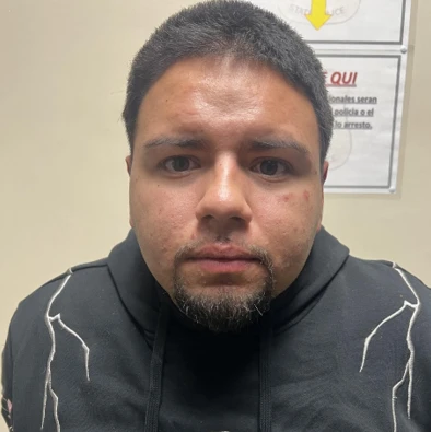 A male stands in front of a tan wall wearing a black hoodie with dark goatee and short dark hair.