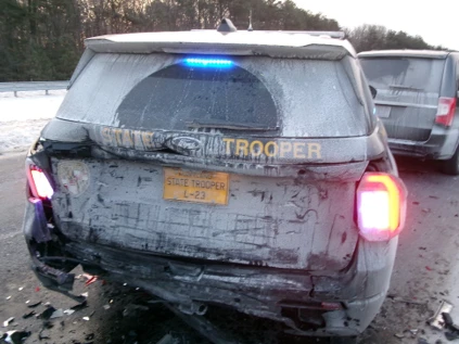 Damaged state trooper vehicle on snowy road with visible dents and broken taillights with another car in the background.