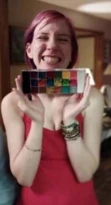 A person with pink hair smiles brightly, holding a vibrant paint palette, wearing a red top and bracelets