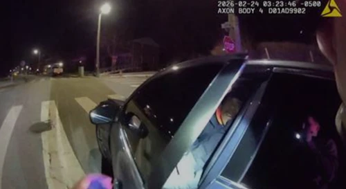 A police body camera footage shows a nighttime traffic stop. A person is seated inside a car at the roadside, illuminated by a flashlight. 