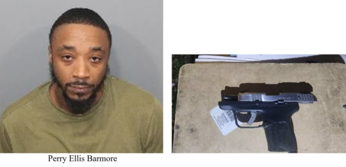 A man wearing a green shirt stands in front of a gray wall, a handgun on the right. 