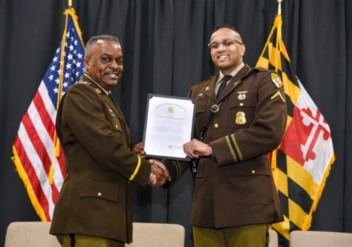 Two uniformed men smiling and shaking hands while holding an award certificate. U.S. and Maryland flags are visible in the background.