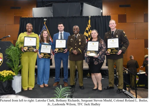 A group of six individuals, two in uniforms and four in civilian attire, stand on a stage smiling and holding certificates.