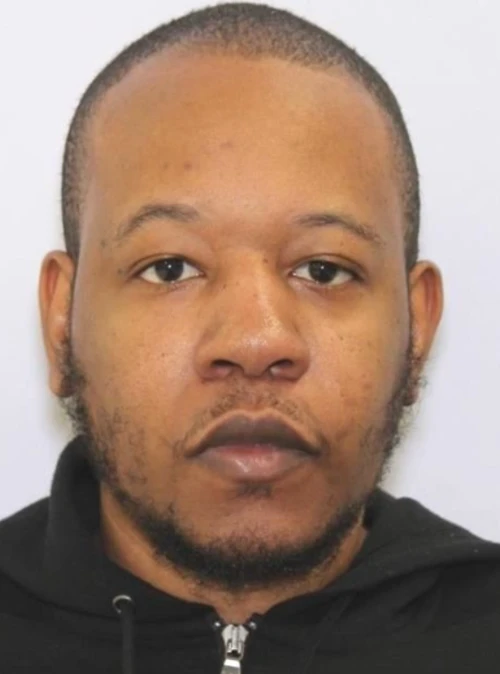 An African American male stands in front of a white background wearing a black hoodie.