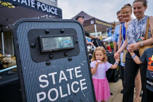 A child peers through a slot in a "State Police" shield at an outdoor event. A girl in a pink skirt eats ice cream nearby, with smiling adults around.