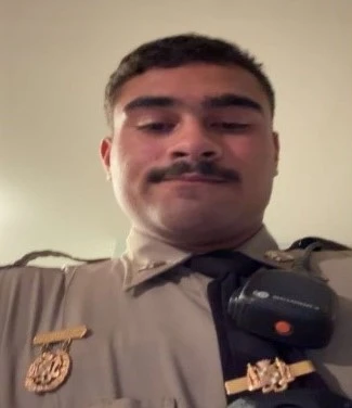 A person in a uniform looks into the camera. They have a badge, radio, and shoulder decorations. 