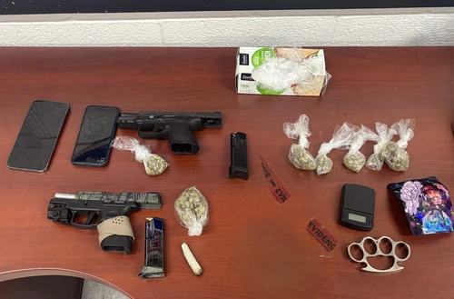 Two smartphones, two pistols, ammunition, plastic bags of cannabis, a scale, brass knuckles, and a colorful packet on a wooden table labeled as evidence.