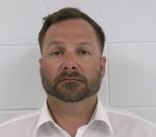 A man stands against a white cinderblock background wearing a white collared shirt with short brown hair and a brown beard.