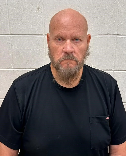 A male standing in front of a white cinderblock wall wearing a black tshirt. 