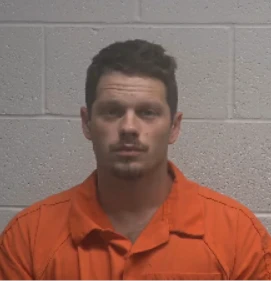 man in an orange prison uniform, standing against a gray cinderblock wall.