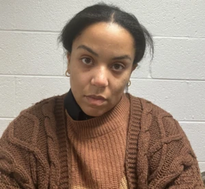 A woman wearing a brown knit sweater layered over a dark collared shirt stands against a white brick wall.