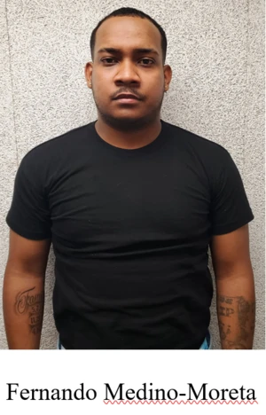 A man stands in front of a gray wall wearing a black tshirt. 