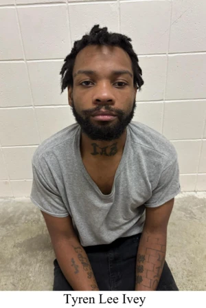 A man with a beard and short dreadlocks is seated against a white tiled wall. He wears a gray T-shirt and black pants, displaying tattoos on his neck and arms.