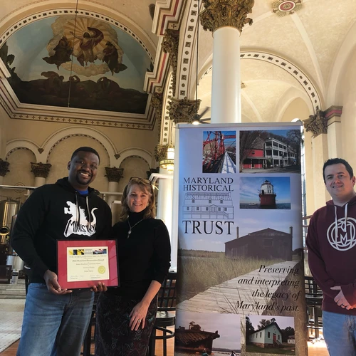Three people standing in a renovated historic interior with religious ceiling murals holding a framed '2022 Maryland Preservation Award' next to a banner for MHT.