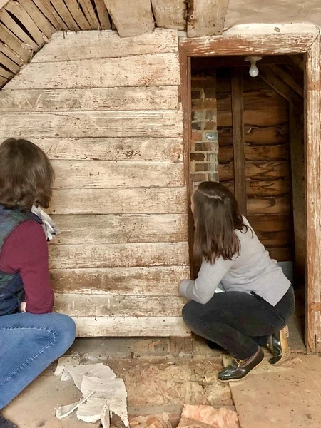 Two preservationists kneel inside a distressed building to inspect a white-washed wooden wall next to a vaulted ceiling with exposed lath.
