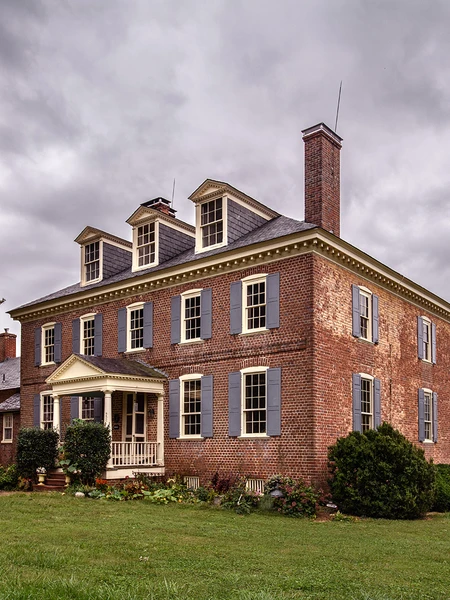 Historic brick house with gray shutters and a gabled porch under a cloudy sky.