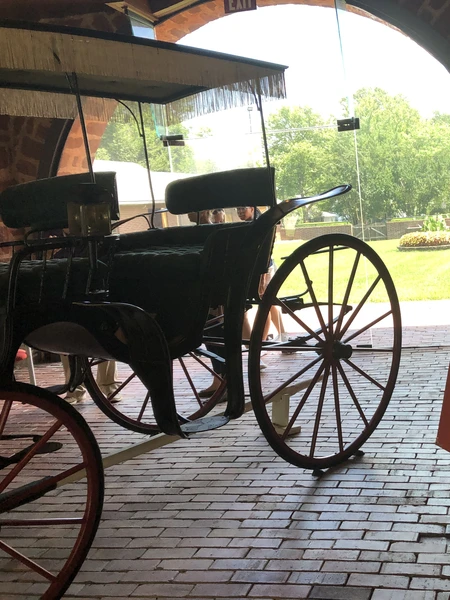 Horse-drawn carriage displayed inside a brick archway with a red “Bel Air Stud Stable” sign and green lawn visible outside.