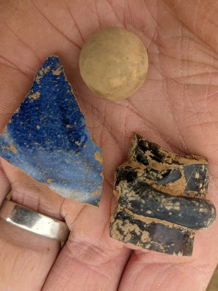 A hand holding a black piece of pottery, a blue fragment, and a small brown ball.