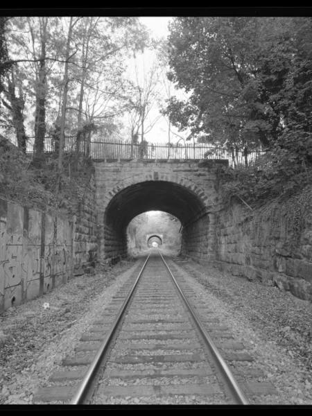 Railway tracks through consecutive stone tunnels, surrounded by graffiti and foliage.