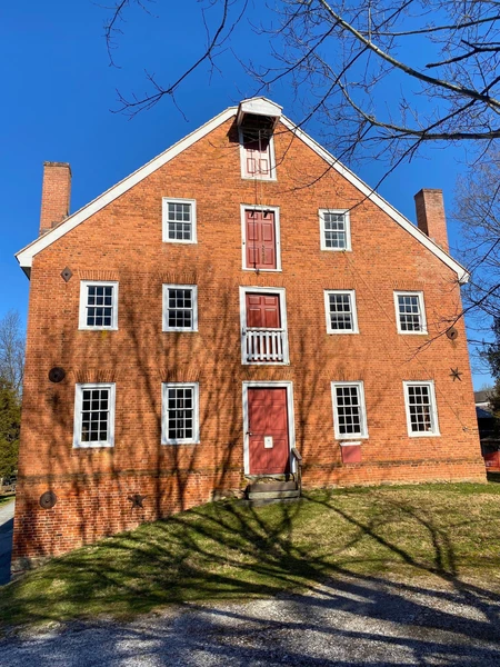 A three-story brick building with red doors and white-trimmed windows, casting tree shadows on its facade under a clear blue sky.