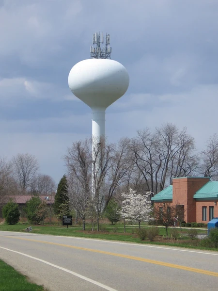 A white water tower with antennas, set against a cloudy sky, near a road and a red-brick building.