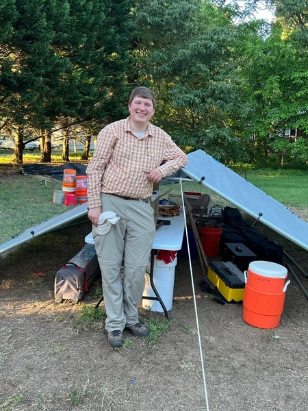 A color photograph of a smiling young man leaning against a temporary tarp shelter at an outdoor archaeological site. Tools and buckets are visible beneath the shelter behind him.